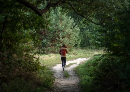 Anonymous distant backpacker with rucksack strolling on trail in woods with lush green trees during trekking in nature on summer day