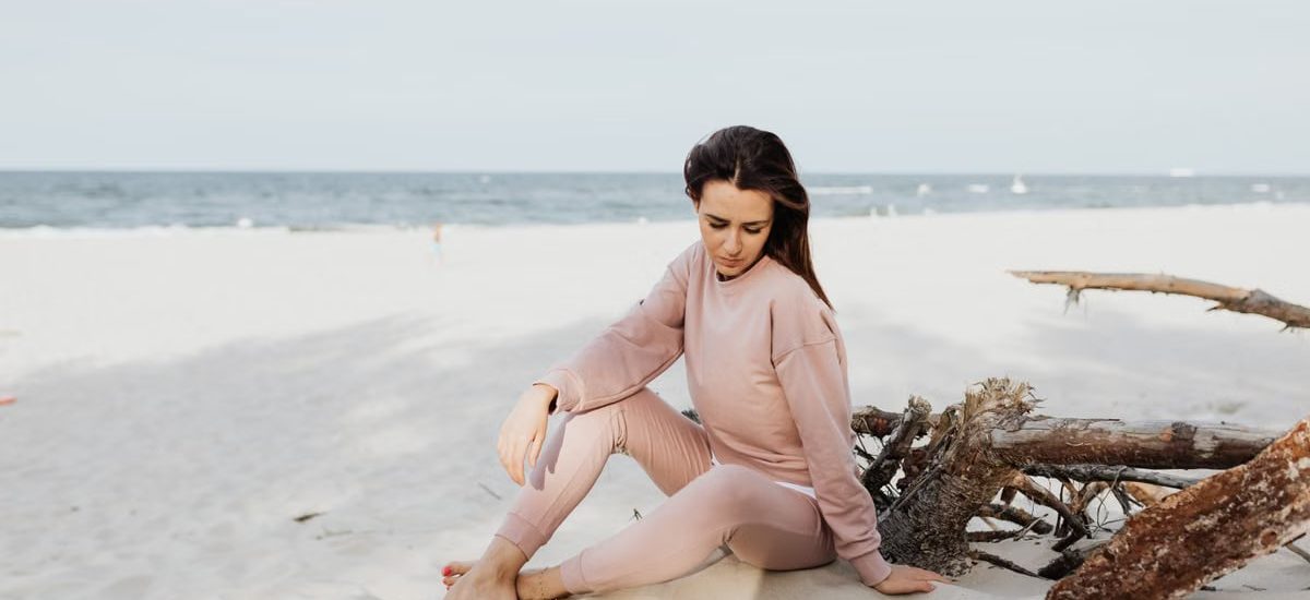 Serene scene of woman pondering alone on a sandy beach in casual pink attire.
